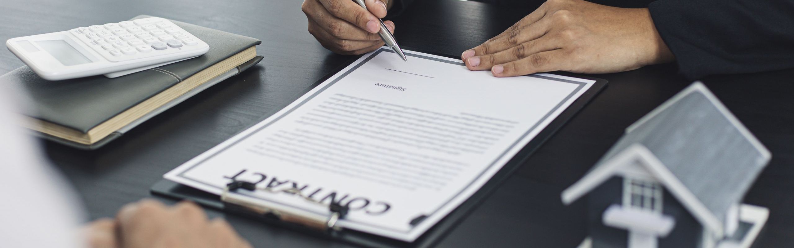 Due persone sedute alla scrivania, una scrive su un contratto, scrivania nera, sulla scrivania &egrave; posata una casetta in miniatura, un contratto e dei faldoni
